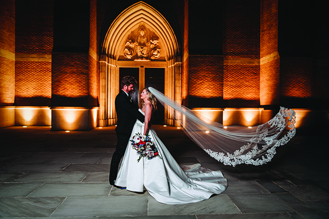 Bride and groom staring at each other while her veil flies in the wind in front of stunning church.