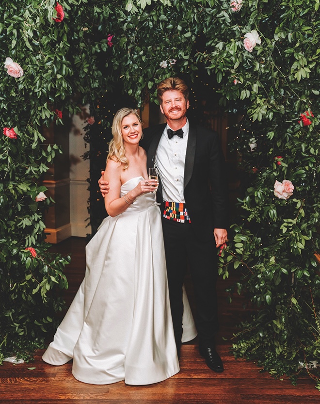 Bride and groom smiling together in front of lush greenery background.