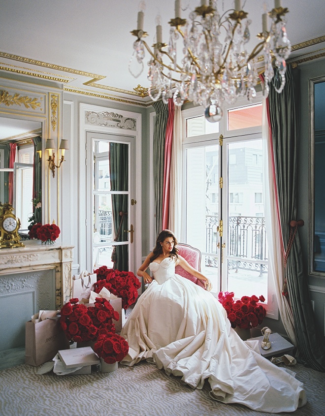 The bride sits in a chair next to several bouquets of red ranunculus and shopping bags in Paris.
