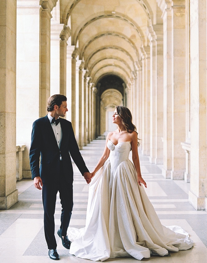 The couple holds hands as they walk under an archway in Paris wedding editorial.