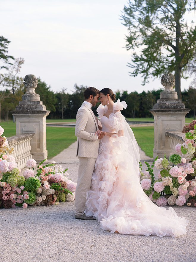The couple hold hands at their outdoor ceremony surrounded by pink and green florals. 