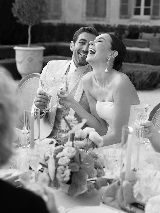 The couple laugh holding Champagne glasses at their reception. 