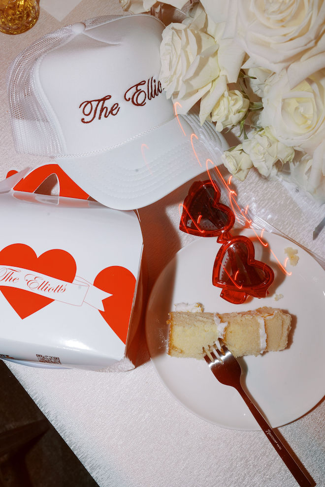Red detailed wedding favors including a white hat, heart sunglasses, and a food box, with a slice of cake next to it.