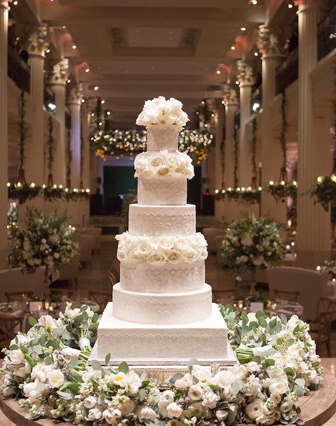 Tall six tier white wedding cake with minor accents and white roses.