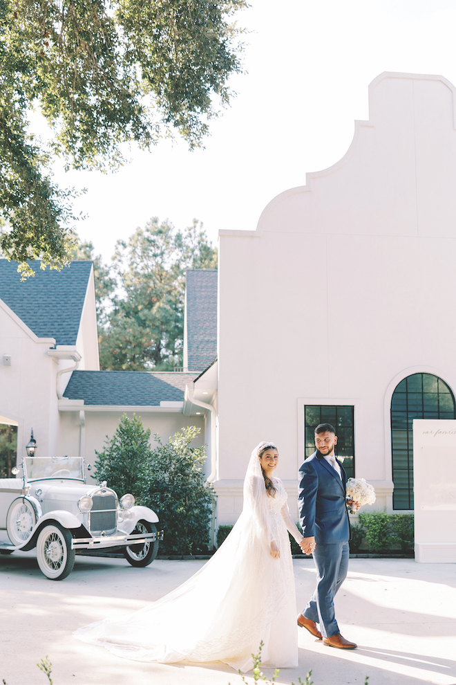 Bride and groom holding hands walking outside of white venue and vintage getaway car behind them.