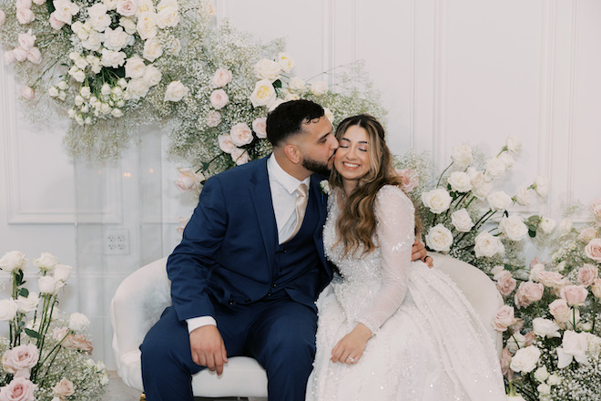 Groom kissing bride on the cheek in front of a floral installation.