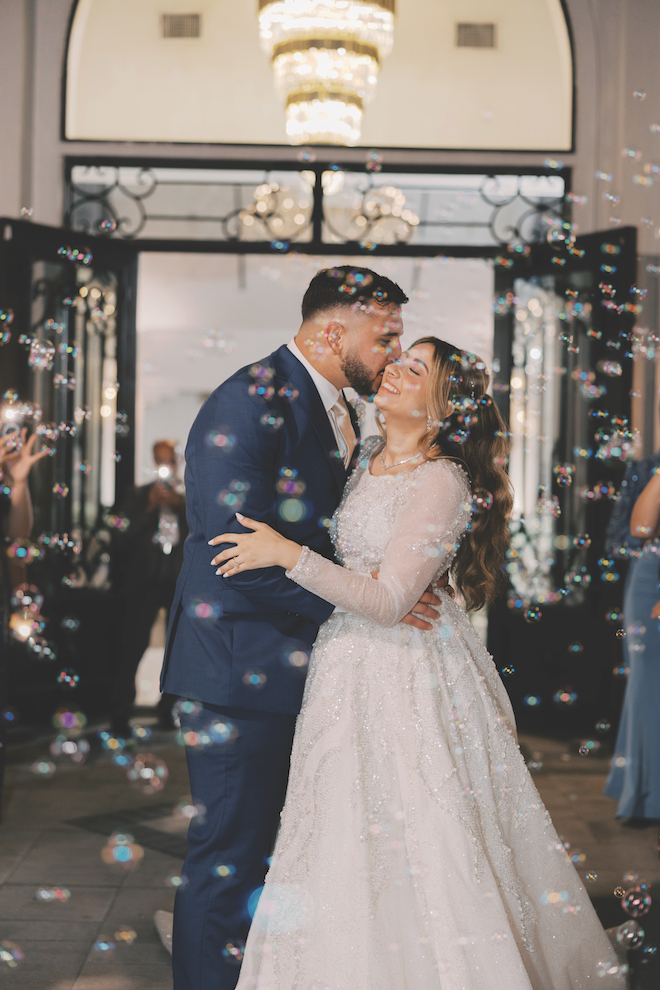 Groom kissing bride on the cheek outside of venue during their grand exit surrounded by guests blowing buubbles.