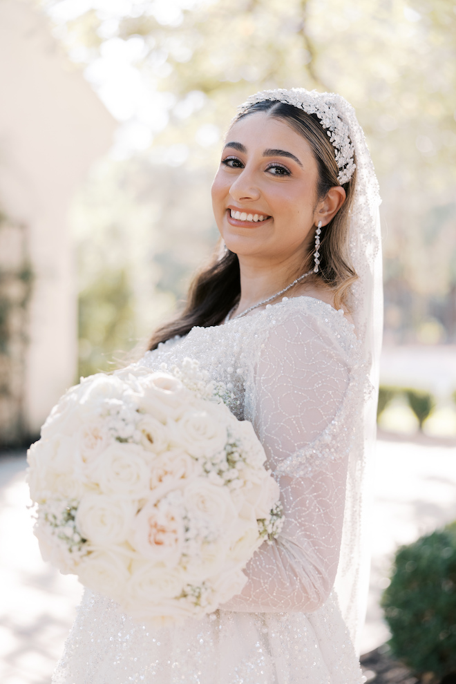 Bride smiling with lacy veil and holding baby's breath and rose bouquet outside of venue.