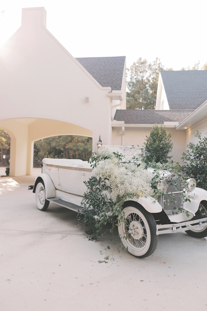 White vintage getaway car with greenery and white florals with baby's breath on it parked outside of venue.