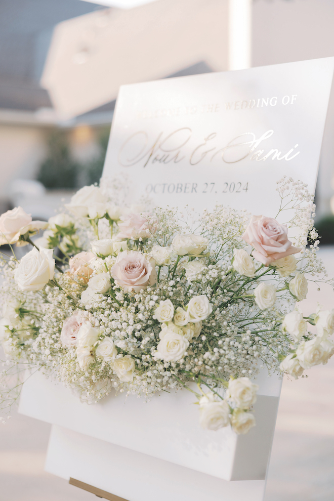 White sign outside of venue with roses and baby's breath that reads, "Welcome to the wedding of Nour & Sami, October 27, 2024"
