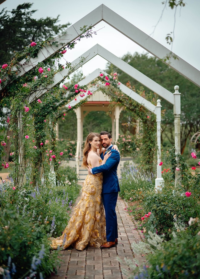 The couple stand on cobble stone in a garden by a gazebo. 