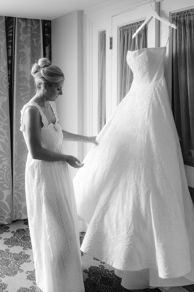 Black and white photo of bride looking at gown hanging up.