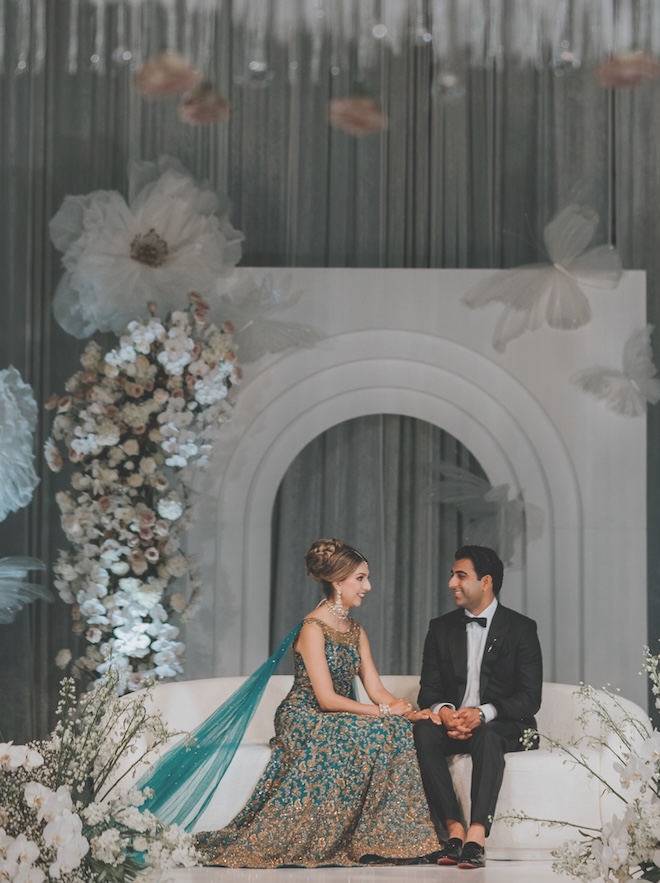 The bride sits on the couch at her wedding reception holding the groom's hand. 