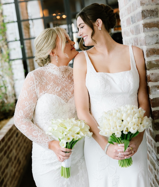 The two brides look into each others eyes and smile while holding their wedding bouquets. 