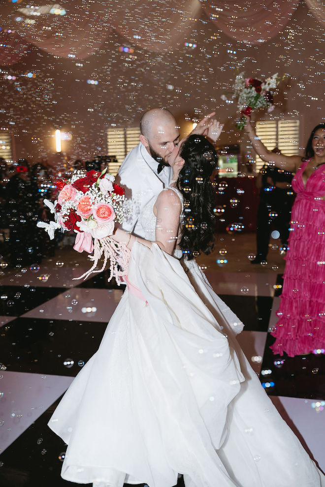 Bride and groom kissing on the dance floor while holding a pink, red and white bouquet.