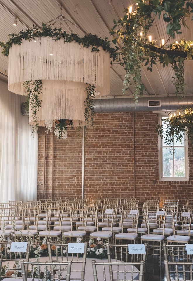 Rope chandelier with greenery above gold wedding guest chairs in front of vintage brick wall.