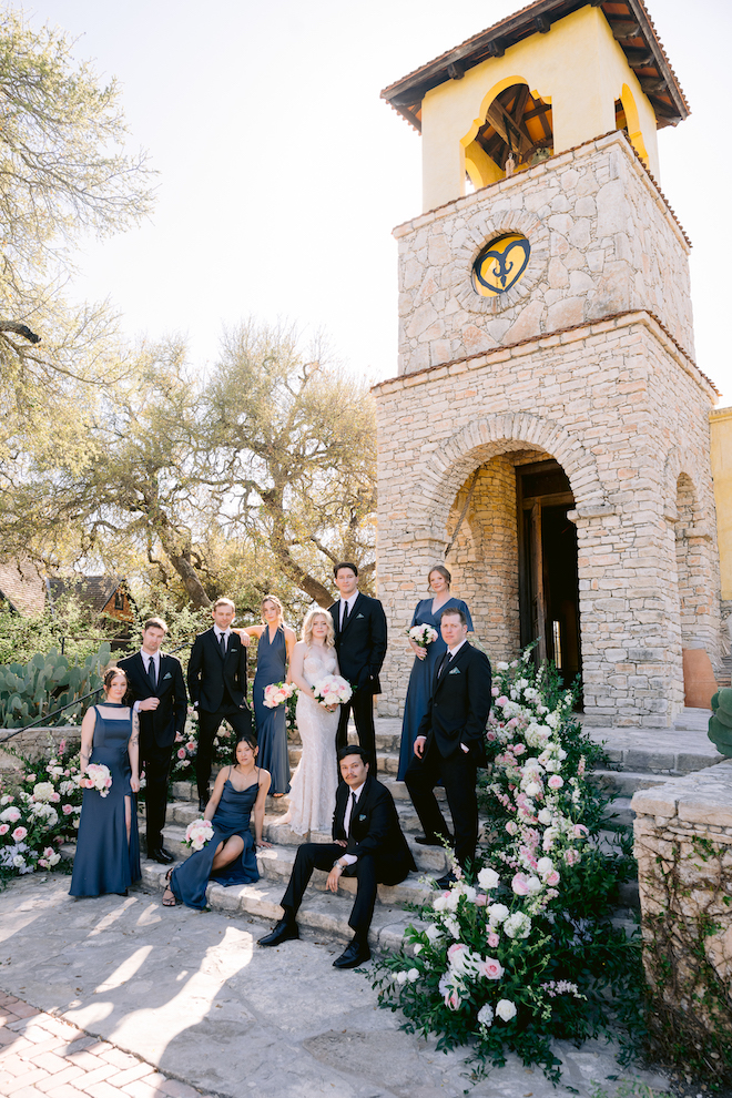 Newlyweds and wedding party in front of the stone church surrounded by greenery and white and pink roses in initimate hill country wedding.