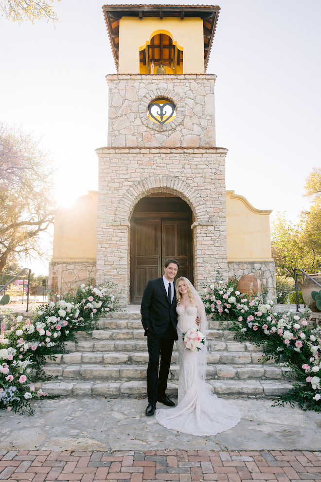 Newlyweds standing in front of stone church together surrounded by greenery with white and pink florals.