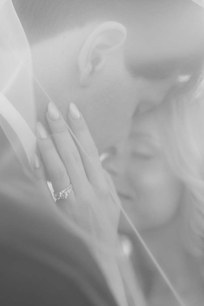 Black and white close up of the newlyweds under her veil showing off her ring.