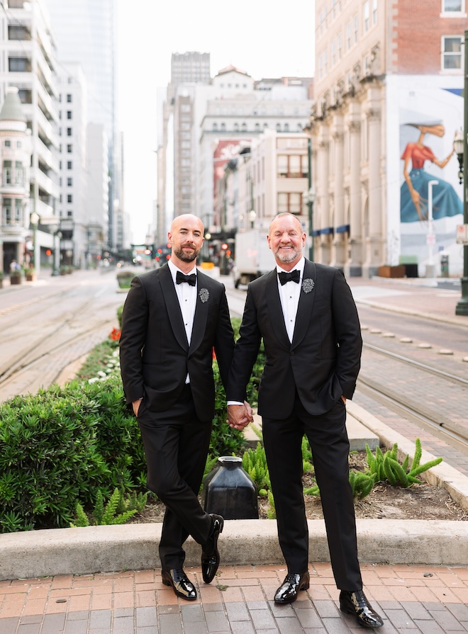 Two groom's hold hands in the center of the city of Houston. 