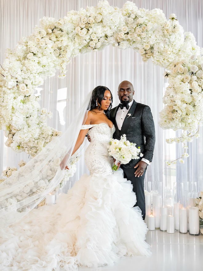 Bride and groom standing together in front of white floral installment for luxury wedding details.