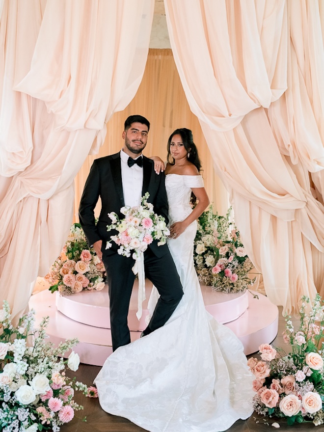 The bride and groom stand in front of the altar where peach drapery and pastel florals decorate the ceremony. 