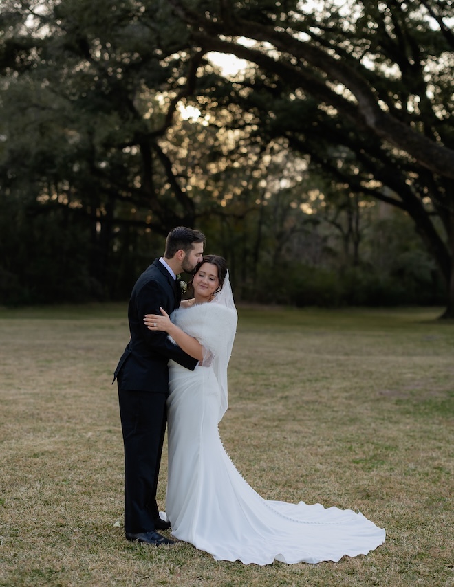 Sammi Sand Photography captures the groom kissing the bride on her head outside in front of a bunch of trees. 