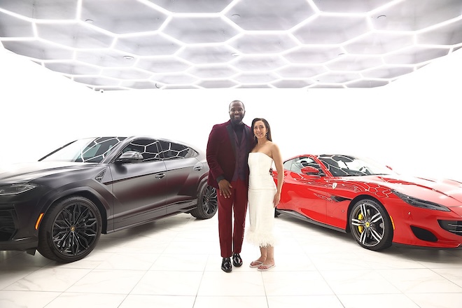 The couple smile beside a Porsche and Ferrari. 