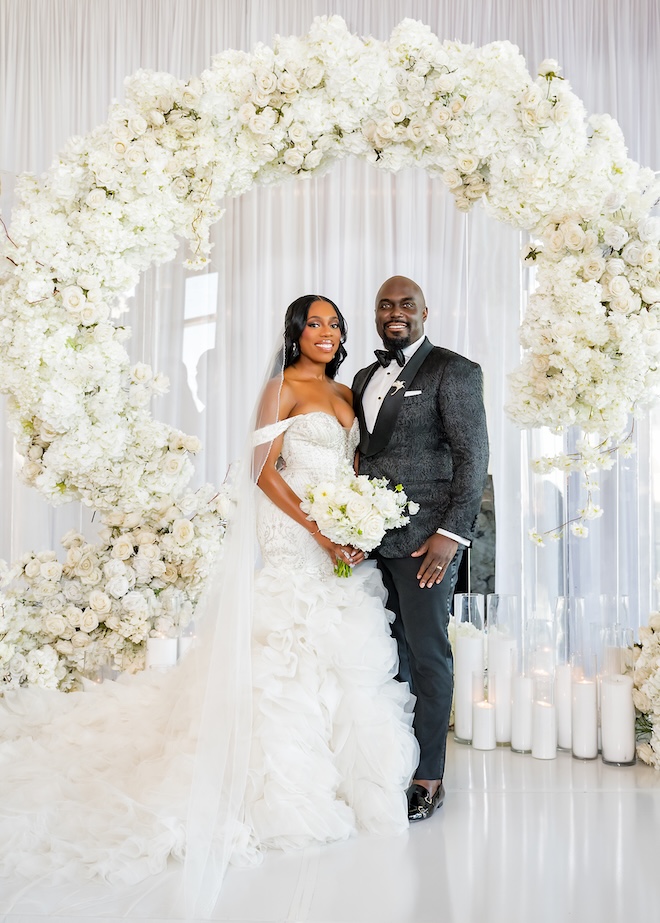 The bride and groom stand in front of an all-white wedding floral arch at their ceremony.