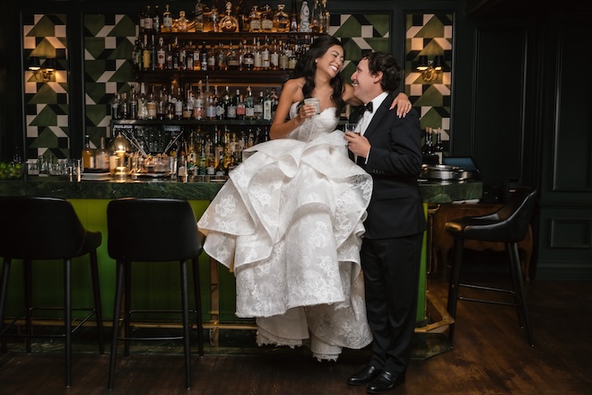 The bride sits on the bar top and shares a cocktail with the groom. 