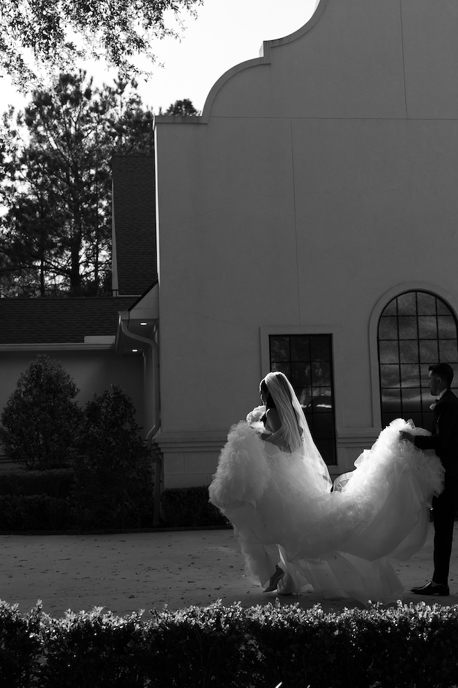 Black and white photo of the groom holding the brides train as they walk towards their garden-inspired wedding.