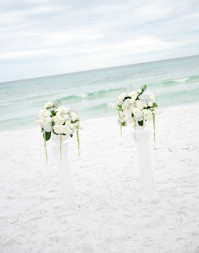 Two white pillars with white and light blue floral bouquets on the beach by the water for the altar.