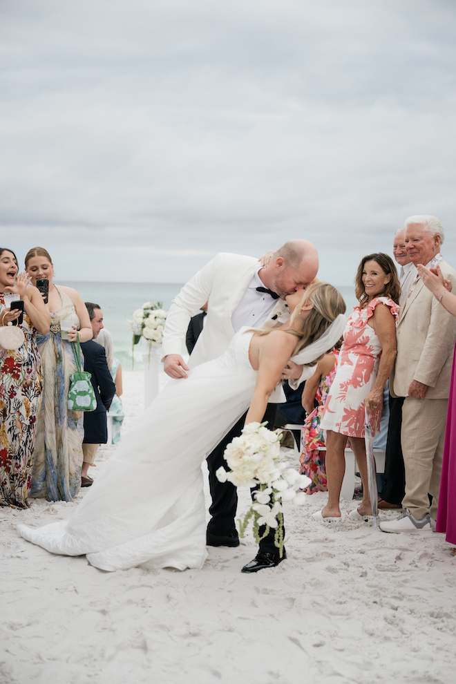 Groom dipping the bride in the middle of the sand aisle and kissing with guests cheering around them.