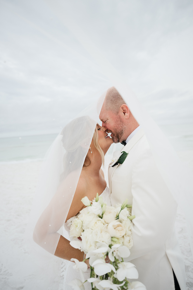 Bride and groom putting their heads together under her veil on the beach.