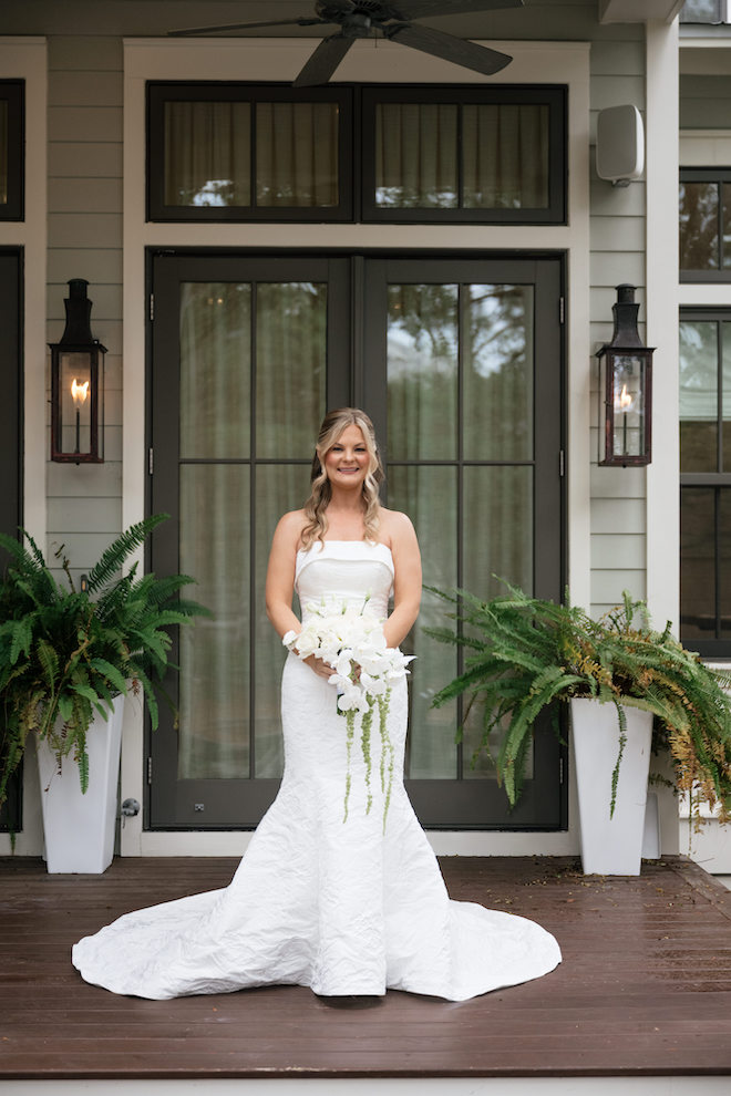 Bride standing outside of french doors in gown holding her bouquet before walking down the aisle for her intimate Seaside wedding.