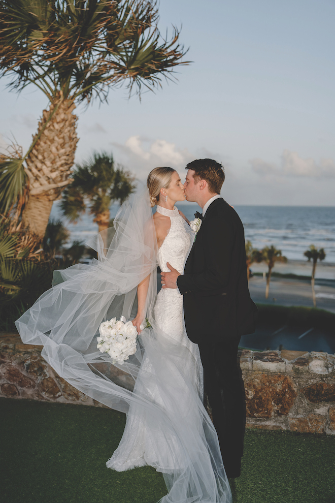 Bride and groom kissing in front of palm trees with the beach behind them.