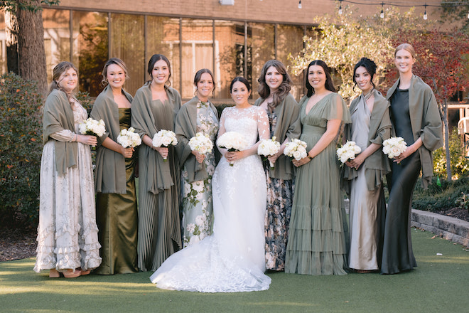 Bride with bridesmaids in shades of green dresses holding white bouquets.