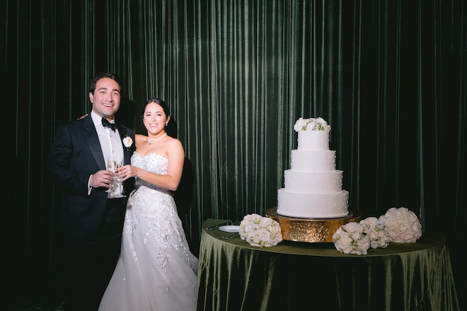 Newlyweds next to 4 tier classic wedding cake in front of green drapery. 