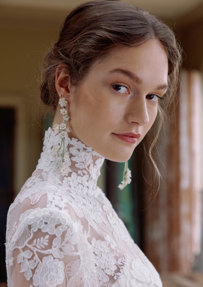 The model looks into the camera wearing a lace wedding dress and white floral dangling earrings.