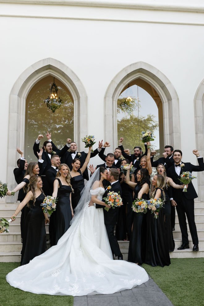 The newlyweds kissing outside of venue with the wedding party cheering around them.
