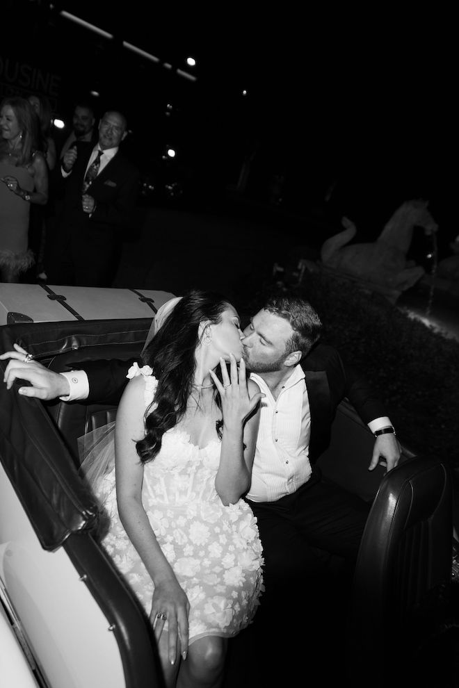 Black-and-white photo of the couple kissing in a getaway car while bride shows off her ring.