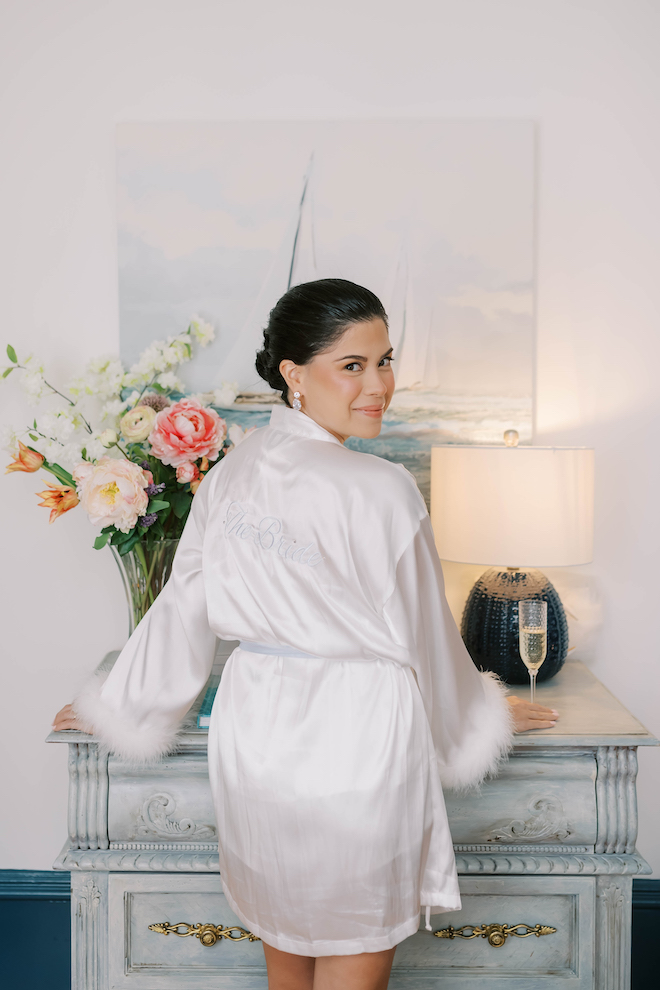Bride in silk bridal robe looking at back at camera with a glass of champagne for bridal pajamas and loungewear.