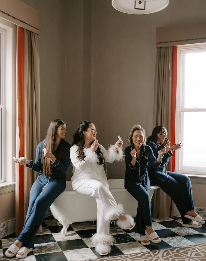 The bride and her bridesmaids sit on the edge of the tub as they put on their makeup. 