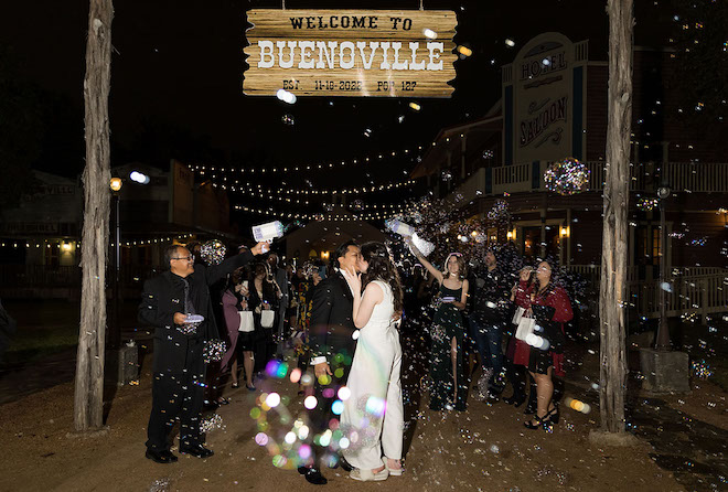 Bride and groom kissing under western sign at night surrounded by bubbles.