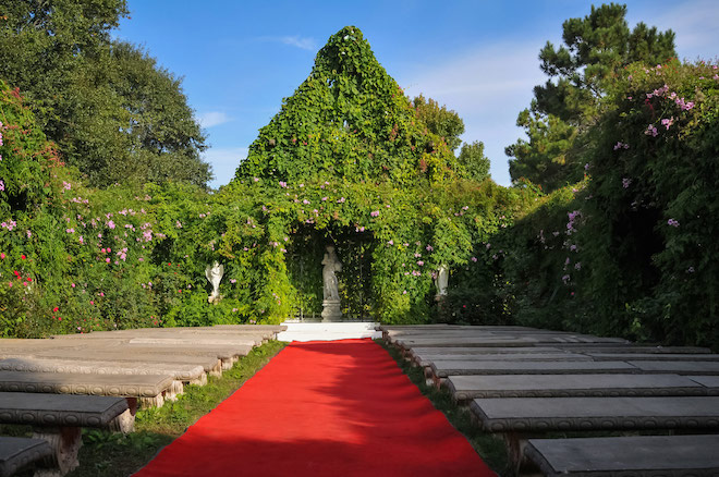 Vine covered chapel with concrete benches with a long red carpet for the aisle.