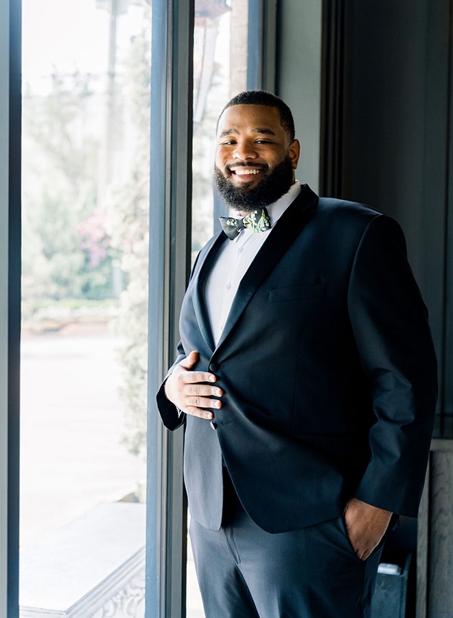 The groom stands next to the window wearing his wedding tuxedo. 
