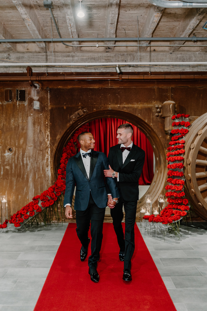 Two grooms outside of vintage bank vault venue adorned in red roses and a red carpet.