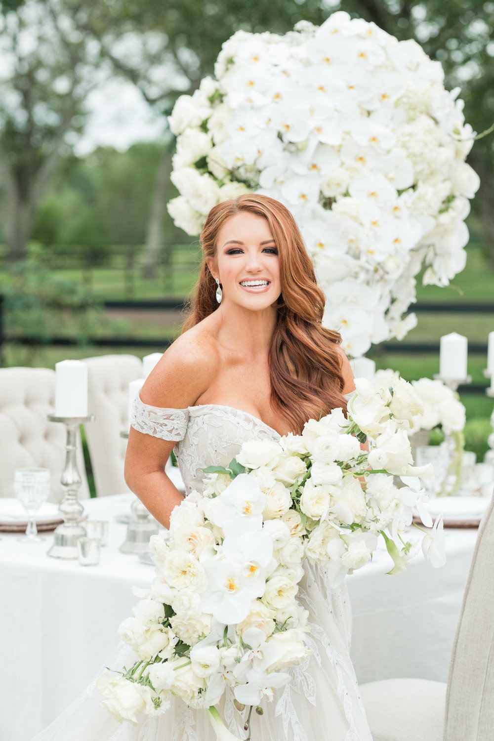 Bride in off the shoulder gown in front of white table setting holding a white bouquet. 